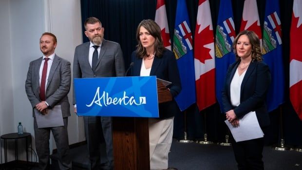 A group of people standing in front of flags, with a woman at the centre speaking at a podium.