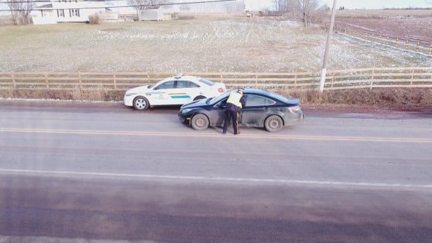 A uniformed member of the RCMP conducts a traffic stop. A person is seen leaning in the window of a black sedan with an RCMP car in the background.