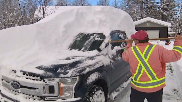 A person in a bright orange jacket uses a broom to clear heavy snow from a parked truck in a snowy driveway.