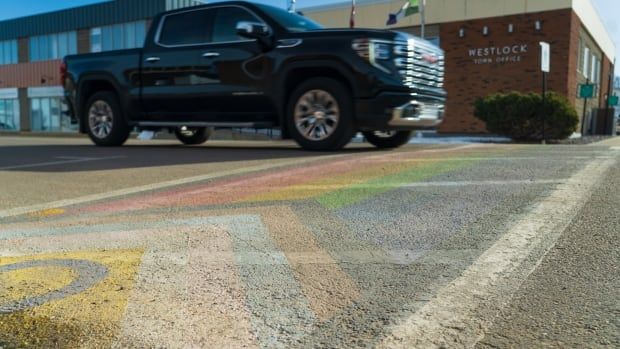 An inclusion rainbow pride flag is painted in front a town hall building. A black truck drives past it.