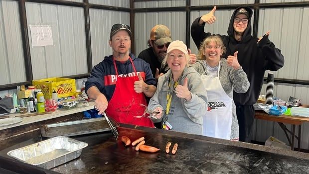 Five people stand behind a big grill at an outdoor event with their thumbs up. A couple of them are holding tongs and spatulas, cooking sausages on the grill.