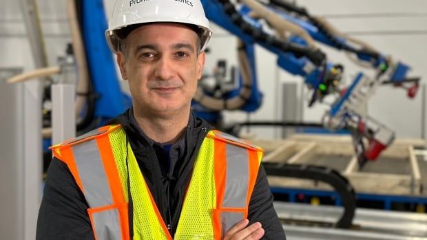 A man wearing a safety vest and white hard hat stands in front of two blue robotic arms working on framing a wall. 