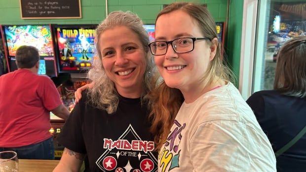 Two woman stand side by side, having their arms around each other. There are pinball machines in the background.