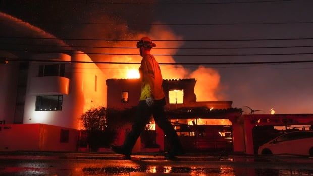 a firefighter walks in front of a burning building at night.