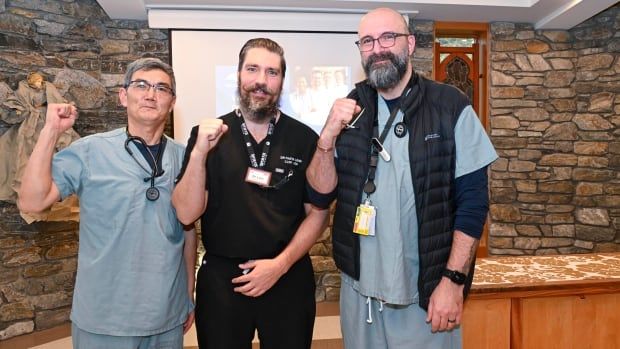 Three men wearing scrubs smile and raise their hands.