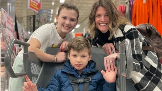 A boy sits in a special cart in the aisle of a store  while two women gather around and smile for the camera