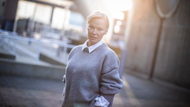 A woman wearing a grey sweater over a white blouse poses with her hands in her pockets.