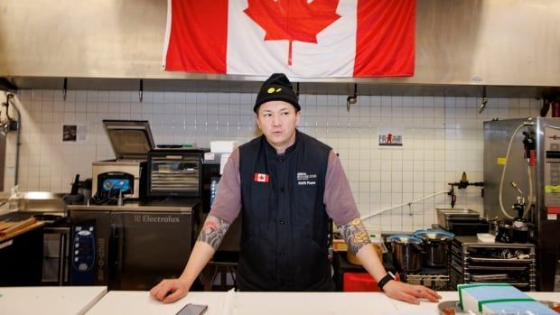 A man is pictured standing behind a table with a giant canadian flag hanging on the wall behind him. he has his arms spread wide and his hands on the table.