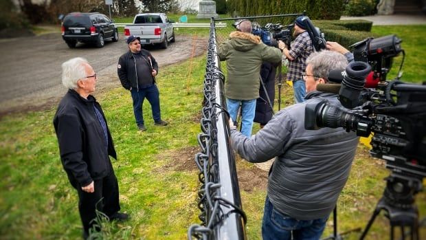 Journalists with TV cameras mass on one side of a black chain-link fence to interview residents on the other side.