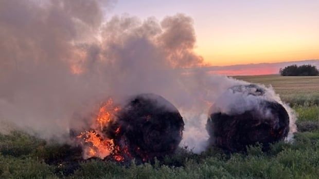 Two large bales of hay burn and give off smoke in a field at dusk.