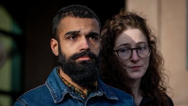 A bearded man is pictured outdoors, with a curly-haired woman to his left in shadow.