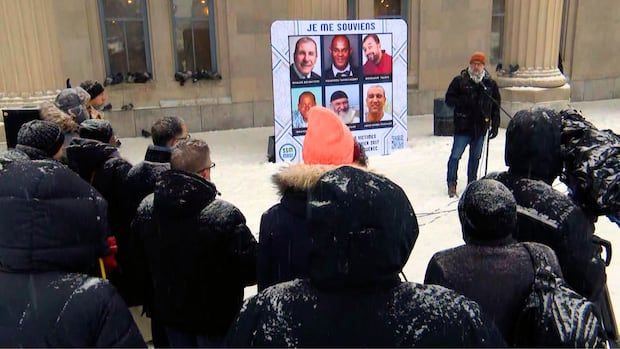 A crowd gathers in a snowy courtyard in front of a man speaking into a microphone with a billboard behind with the faces of six people