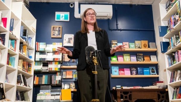 A woman in a blazer speaks into a microphone. Shelves display rows of books in the background.