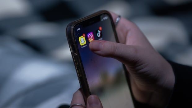 A person wearing multiple silver rings holds a smartphone with their thumb poised over the Snapchat app. 