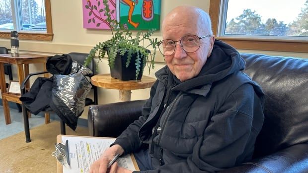 An elderly man sits in a chair wearing a vest, with a clipboard in his lap and a colourful piece of Indigenous art on the wall behind him.