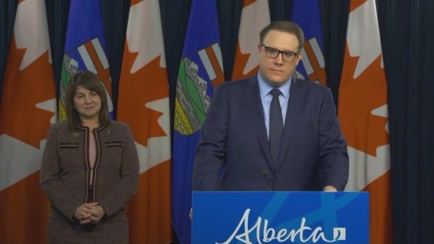 A woman in a brown suit stands next to a man in a blue suit at a podium in front of the flags of Canada and Alberta.