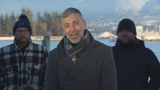 A man in a grey coat with a scarf wrapped around his next, speaks at a news conference.