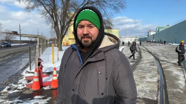 A man standing outside with a snowy background wearing a winter coat and winter hat looks into the camera.