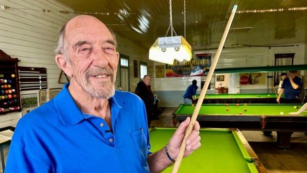 A man in a bright blue shirt stands with a pool cue in front of a large green pool table in a rustic pool hall.