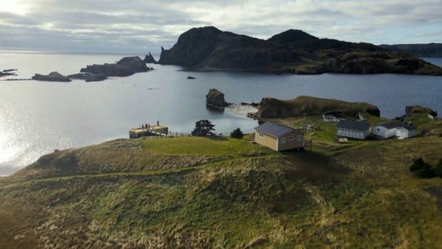 A panoramic photo captured with a drone of a grassy island with a few homes on it. A few people are standing on the island's wharf.