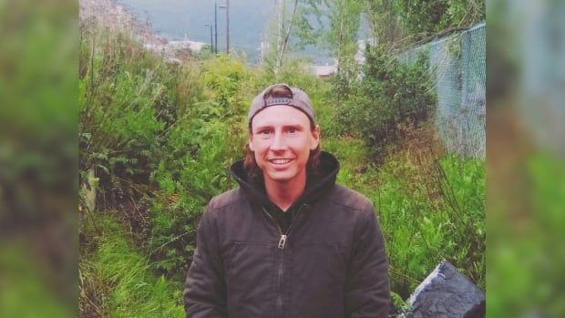 A young man is seen outside, dressed in a hoodie, zip-up jacket and backwards baseball cap. 