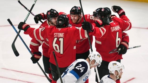 A group of ice hockey players wrap their arms around each other in celebration as two opposing players skate away.