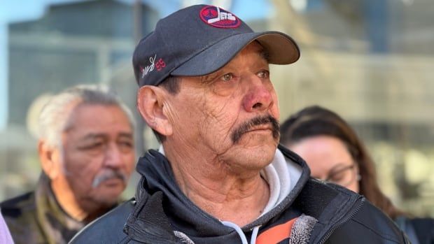 A teary-eyed man wearing a baseball cap and hooded sweatshirt is seen in a close crop, showing his head and shoulders.