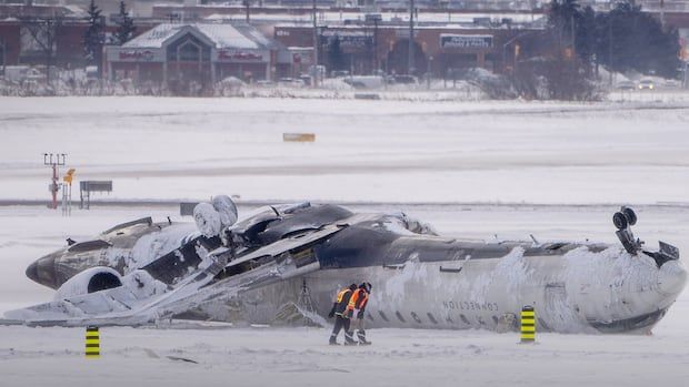 People in safety gear walk toward a badly plane that's upside down.