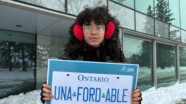 A person in a winter coat stands outside holding a sign that looks like a licence plate and says "Una-FORD-able."