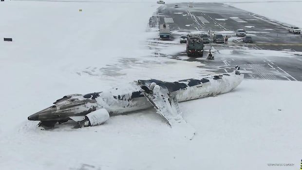 Burned remnants of a crashed plane on a snowy runway with emergency vehicles in the background