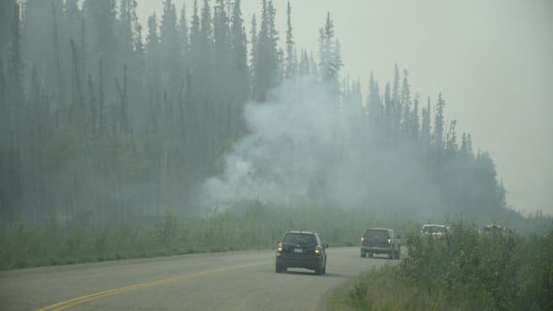 A line of cars drive through wildfire smoke on a rural highway.