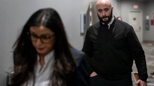 A man in a dark jacket, white shirt and dark tie walks behind a woman on his way out of a hearing.