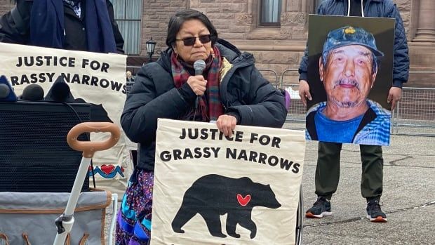 A person in a wheelchair is seen sitting outside and speaking into a microphone. They are holding a sign that says "Justice for Grassy Narrows."