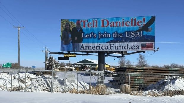 A billboard overlooks a highway on a snowy day. It features a picture of U.S President Donald Trump and Alberta Premier Danielle Smith smiling as an eagle flies by in the background. In big letters, it reads: "Tell Danielle! Let's join the USA! AmericaFund.ca"  An American flag is in the lower-right corner. 