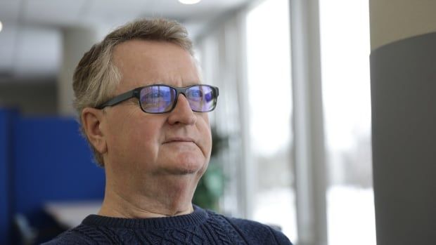 A grey-haired man in glasses and a dark blue sweater looks out the window.