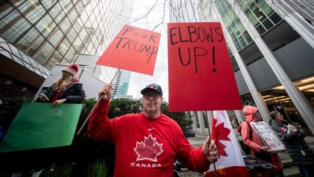 A man in a Canadian hockey jersey and Canada hat holds up two red signs, one reading "F**k Trump," the other reading "Elbows up!" as other people holding signs stand beside him on a street outside tall office buildings. 