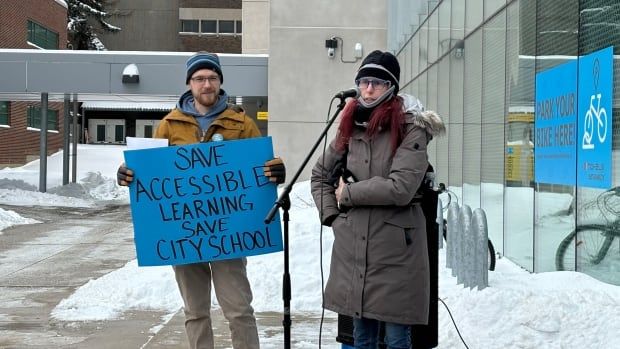 A man holding a sign that reads 'Save accessible learning. Save City School,' next to a woman speaking at a mic.