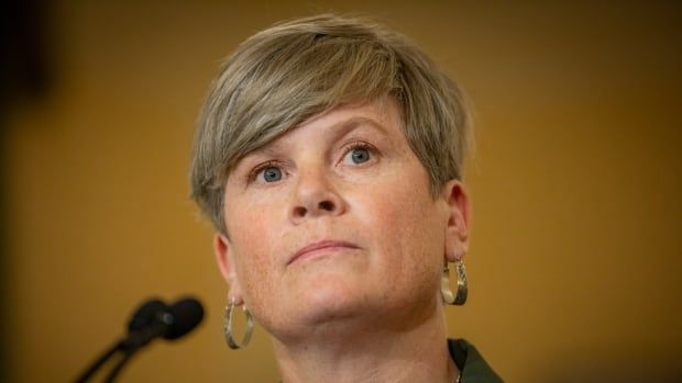 A white woman with short hair and hooped earrings is seen at a mic.