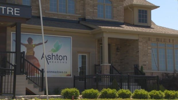 A shot of a beige brick home with a large sign that reads "Ashton Meadows" with an image of a child on their parents shoulders.