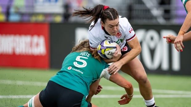 A woman in a green rugby uniform grabs the leg of a woman in a white rugby uniform who is clutching a rugby ball 