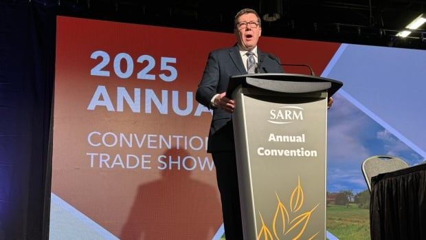 A man in a black suit stands at a lectern with a large screen behind him. The screen reads "2025 Annual Convent and Trade Show."