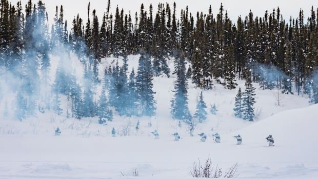 Smoke across a snow covered valley with conifer trees in the background. Soldiers dressed in white kneel with rifles drawn.