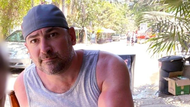 A man in a baseball cap wearing a cut-off shirt sits with palm fronds visible in the background. 