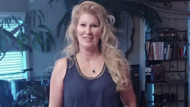 A woman with long blonde hair poses in a living room in a tank top. 