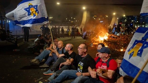 A line of men sit on the ground with linked arms, some holding Israeli flags. A fire burns in the road behind them. 