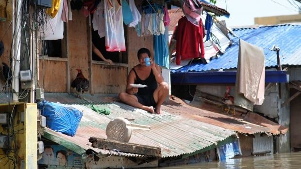 A person sits on a corrugated roof and eats as murky water has risen just below the roof level. Above the person, clothes hang out on a line.