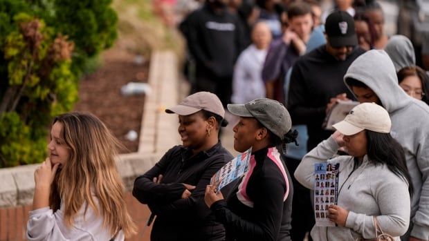 Dozens of people stand in a line up outside. Trees are in the background.