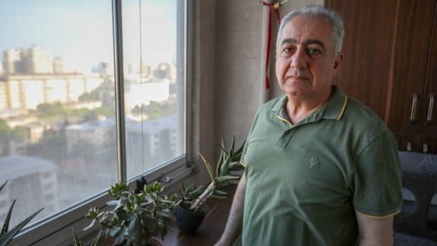 A frowning gray haired man stands by a windowsill covered in plants