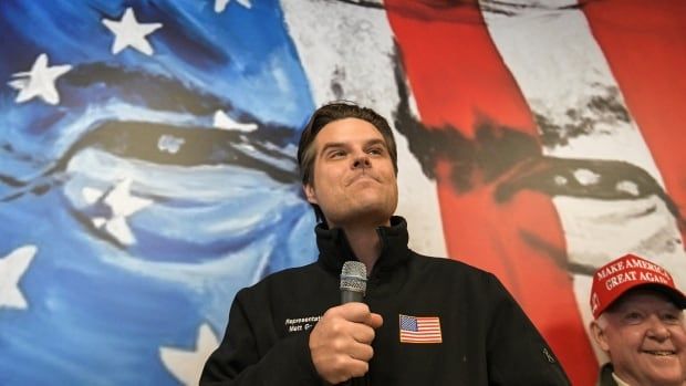 A man in a windbreaker stands on stage, holding a microphone. Behind him is a backdrop of a frowning face, overlaid with red, white and blue stripes.