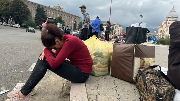 A woman wearing a red top and black pants rests her head on her crossed arms as she sits on a curb with her belongings.